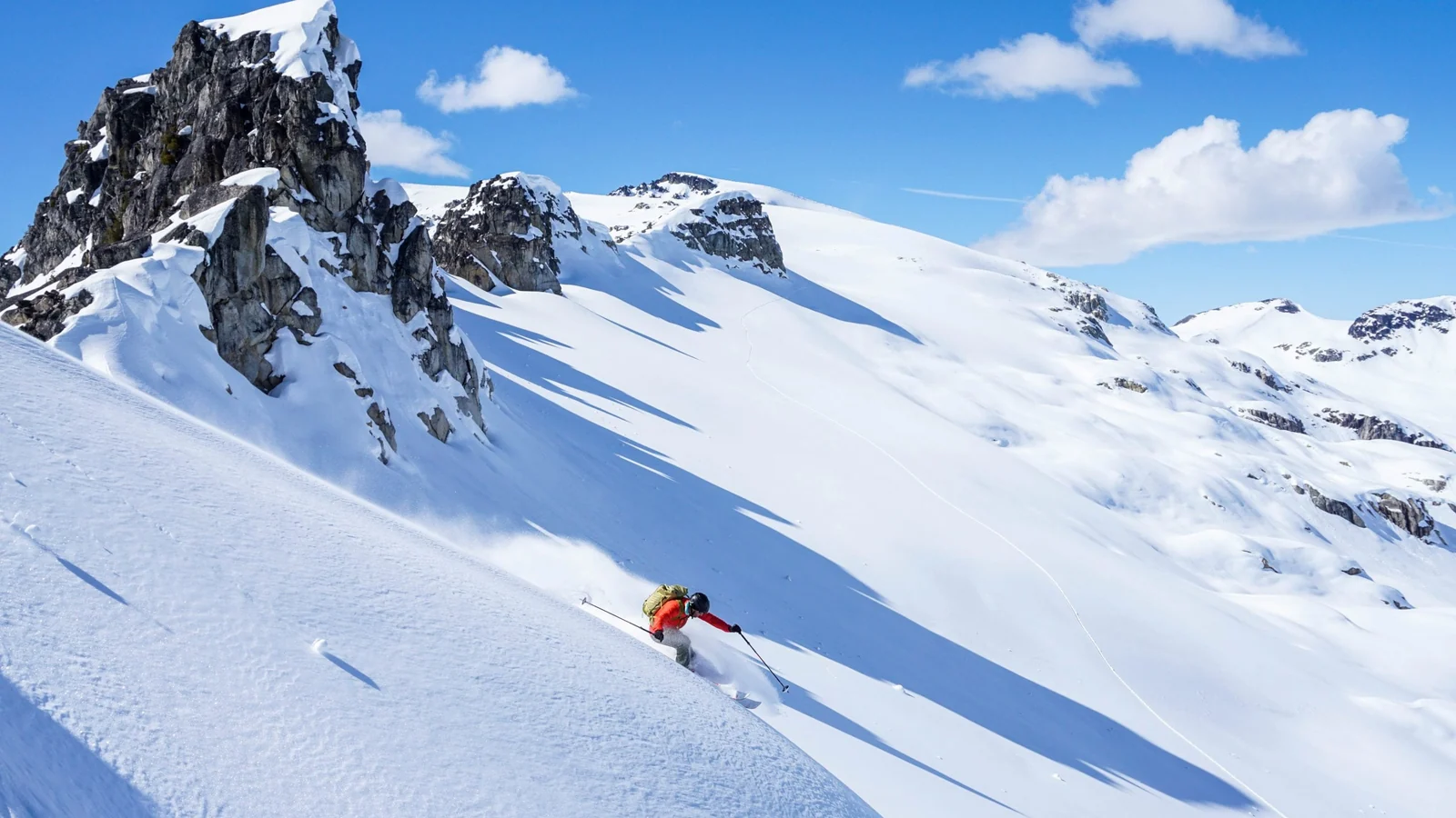 A skier carving down a sunlit face below a rocky summit